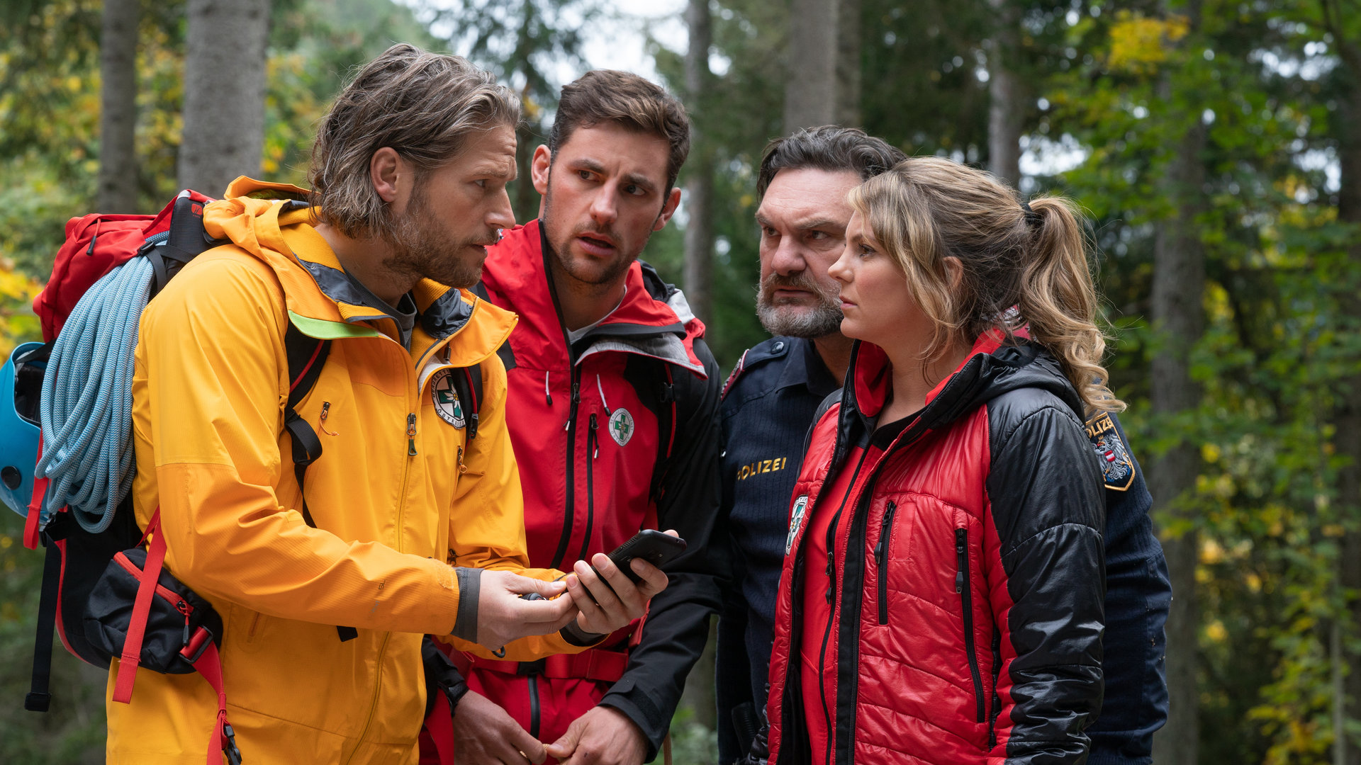 "Die Bergretter: Vierzehn Stufen": Im Wald stehen die Bergretter Markus (Sebastian Ströbel), Simon (Ferdinand Seebacher) und Katharina (Luise Bähr) sowie Polizist Gert (Martin Leutgeb) beisammen und blicken sich fragend an. Markus hält ein Handy in der Hand.