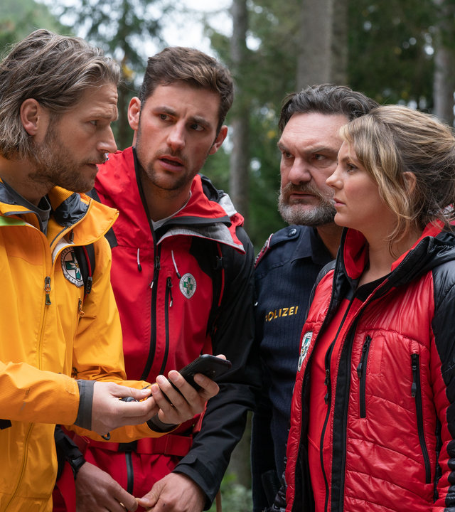 "Die Bergretter: Vierzehn Stufen": Im Wald stehen die Bergretter Markus (Sebastian Ströbel), Simon (Ferdinand Seebacher) und Katharina (Luise Bähr) sowie Polizist Gert (Martin Leutgeb) beisammen und blicken sich fragend an. Markus hält ein Handy in der Hand.