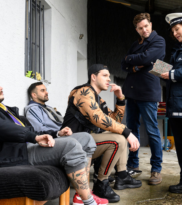 "SOKO Stuttgart – Nachts im Museum": Jo Stoll (Peter Ketnath) und Sibylle Beyer (Bärbel Stolz) stehen auf einem überdachten Vorbau und befragen den Rapper Marc Jessner (Bastian Hagen). Links und rechts von ihm sitzen seine Rapper-Kollegen (Komparsen). Alle drei Tragen Hip Hop-Kleidung.
