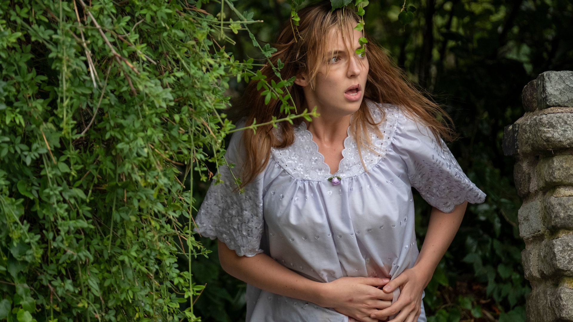 "Killing Eve - Das ist es, was du kriegst": Villanelle (Jodie Comer) steht in einem Nachthemd vor einer Hecke und schaut panisch zur Seite, während sie mit beiden Händen eine blutende Wunde an ihrem Bauch abdeckt.
