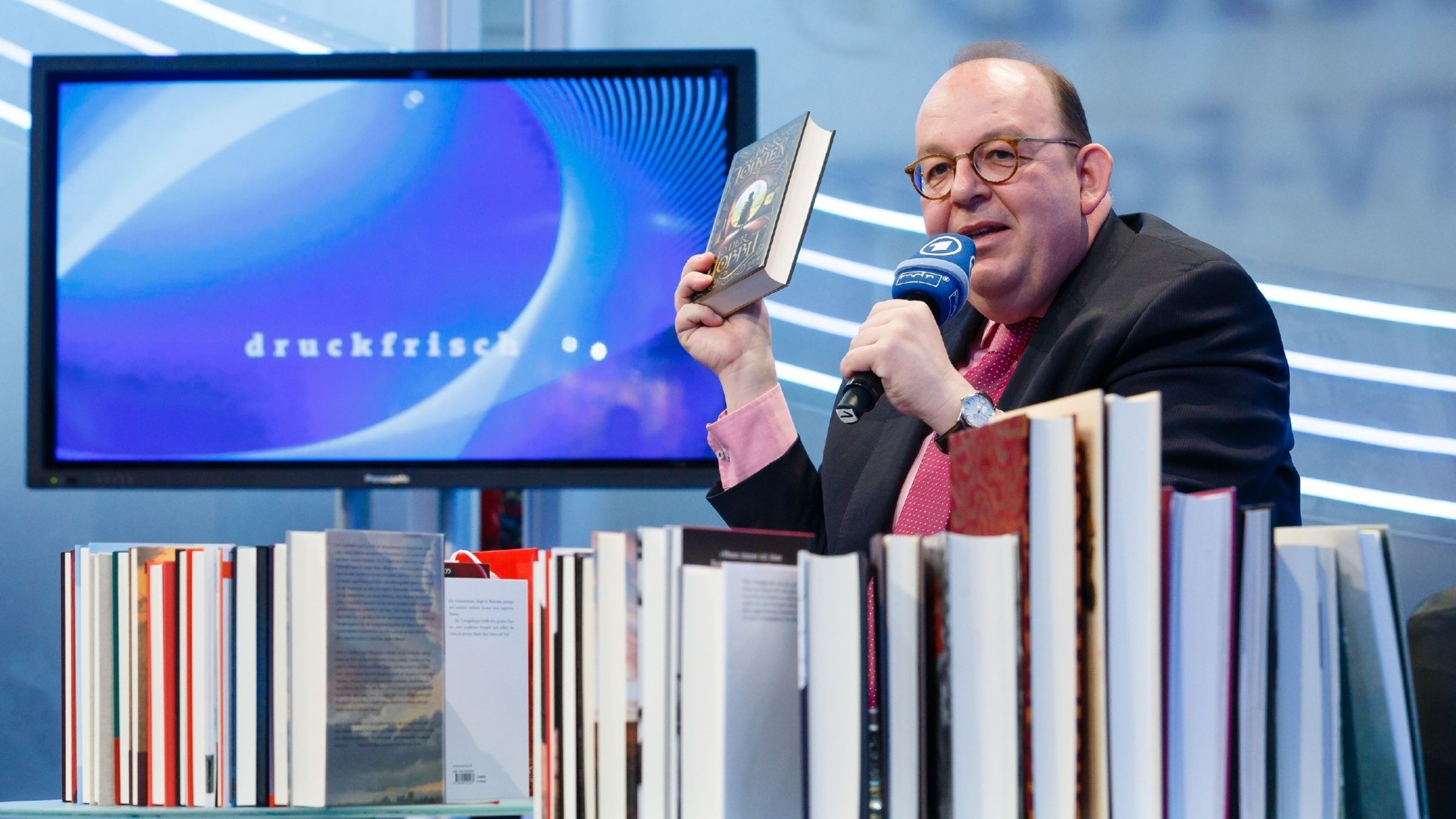 "Druckfrisch": Denis Scheck sitzt hinter einer Reihe von Büchern. Er hält in seiner rechten Hand ein Buch hoch und in der linken Hand ein Mikrofon vor seinen Mund. Hinter ihm steht ein Bildschrim auf dem das Wort "druckfrisch" in kleinen Buchstaben zu lesen ist.