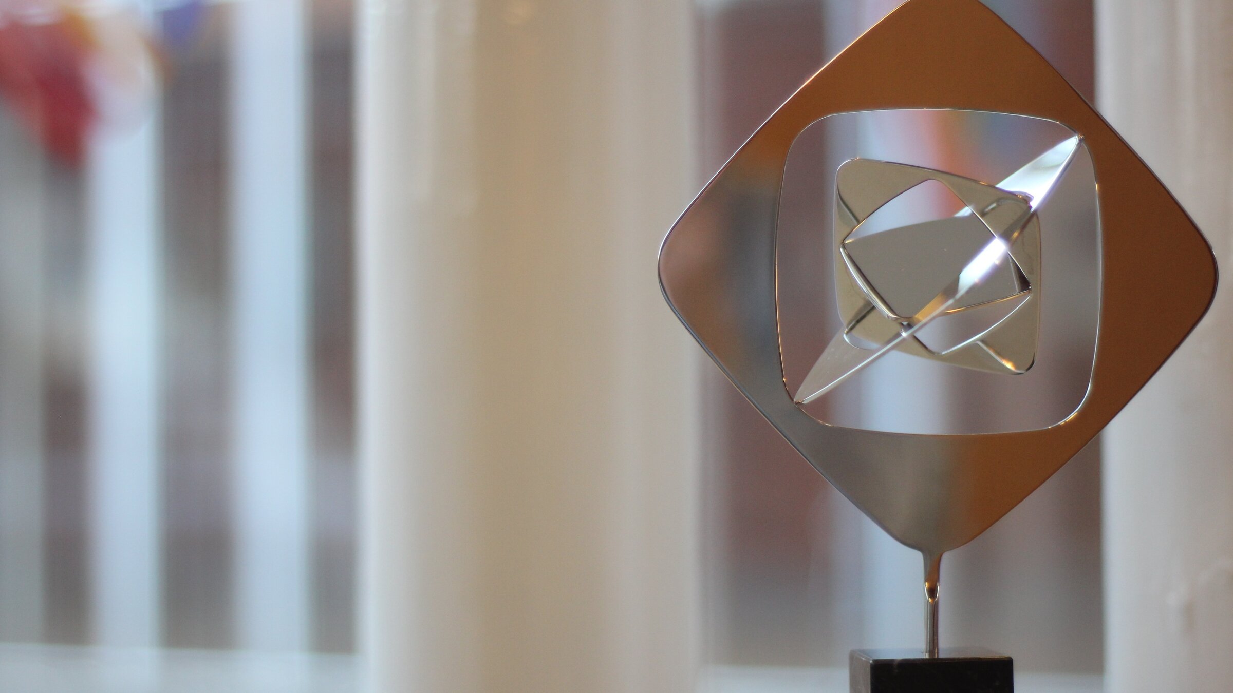 "Verleihung des 62. Grimme-Preises 2026": Das Bild zeigt eine moderne, abstrakte Skulptur aus poliertem Metall, die auf einem schwarzen Sockel steht, vor einem unscharfen Hintergrund eines Fensters mit weißen Vertikalstreben. Die Skulptur hat eine rautenförmige Form und besteht aus mehreren sich überschneidenden, geometrischen Elementen, die einen dreidimensionalen Effekt erzeugen. Die Oberfläche des Metalls ist hochglanzpoliert und reflektiert das Umgebungslicht. Die allgemeine Atmosphäre des Bildes ist ruhig, minimalistisch und modern.