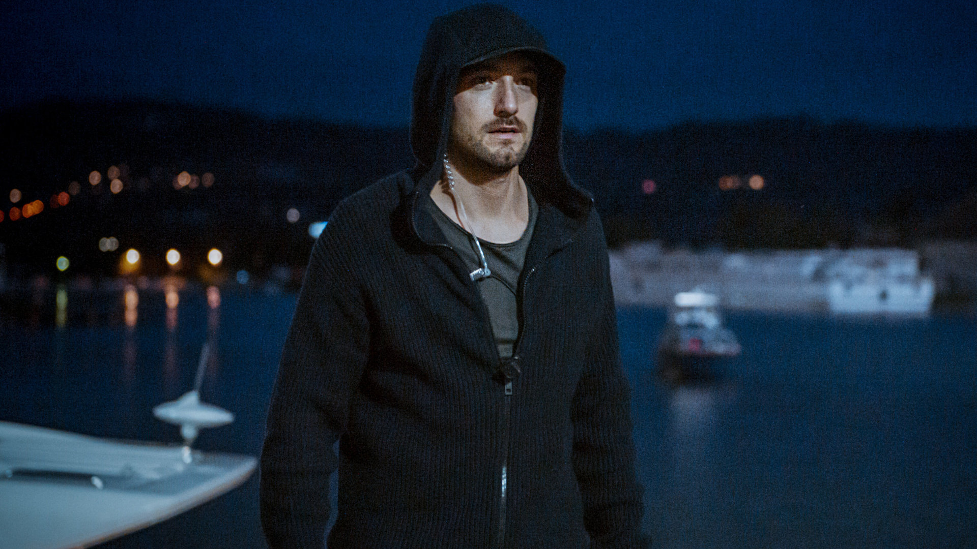 "SOKO Linz - Grenzverschiebung": Im dunklen Kapuzenpullover steht Ben Halberg (Daniel Gawlowski) auf einem Motorboot und schaut vor sich. Hinter ihm sind Boot und Schiff zu erkennen, die auf dem Fluss treiben.