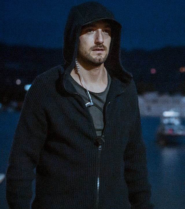 "SOKO Linz - Grenzverschiebung": Im dunklen Kapuzenpullover steht Ben Halberg (Daniel Gawlowski) auf einem Motorboot und schaut vor sich. Hinter ihm sind Boot und Schiff zu erkennen, die auf dem Fluss treiben.