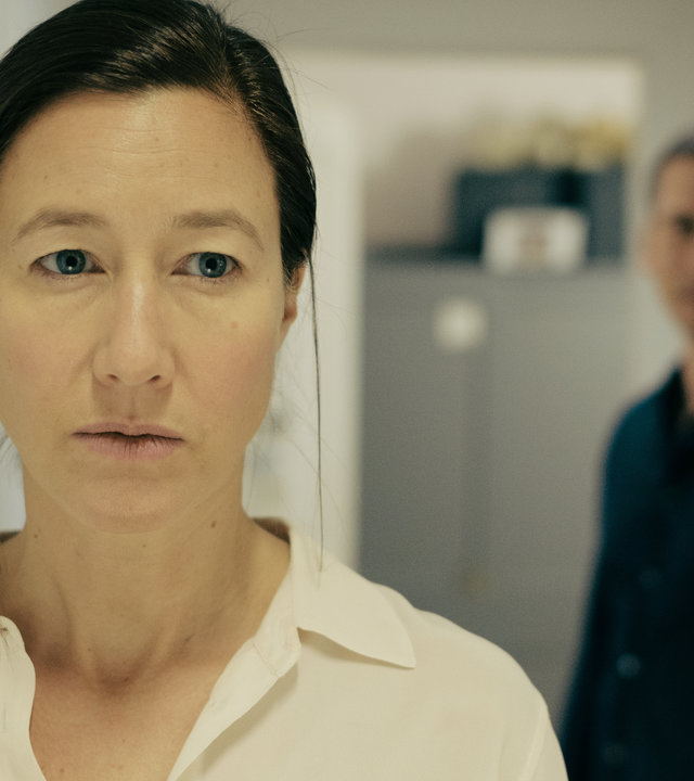 "Am Anschlag - Die Macht der Kränkung - SARAH": Sarah (Johanna Wokalek) sieht in die Ferne. Sie trägt eine Bluse. Im Hintergrund ist Erik (Till Firit) zu sehen.