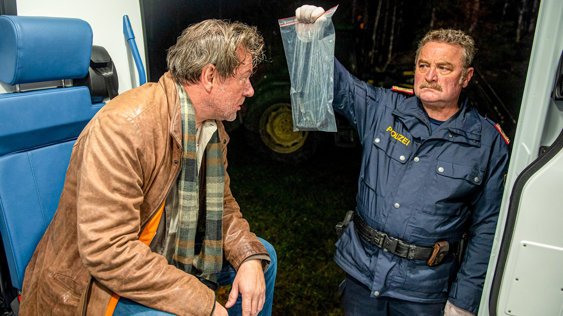 "SOKO Kitzbühel - Zwei Fliegen": Walter Maisinger (Norman Hacker) sitzt in einem Fahrzeug mit offener Seitentür. An der geöffneten Tür steht Kroisleitner (Ferry Öllinger), der ihm eine Waffe in einer Plastiktüte zeigt.