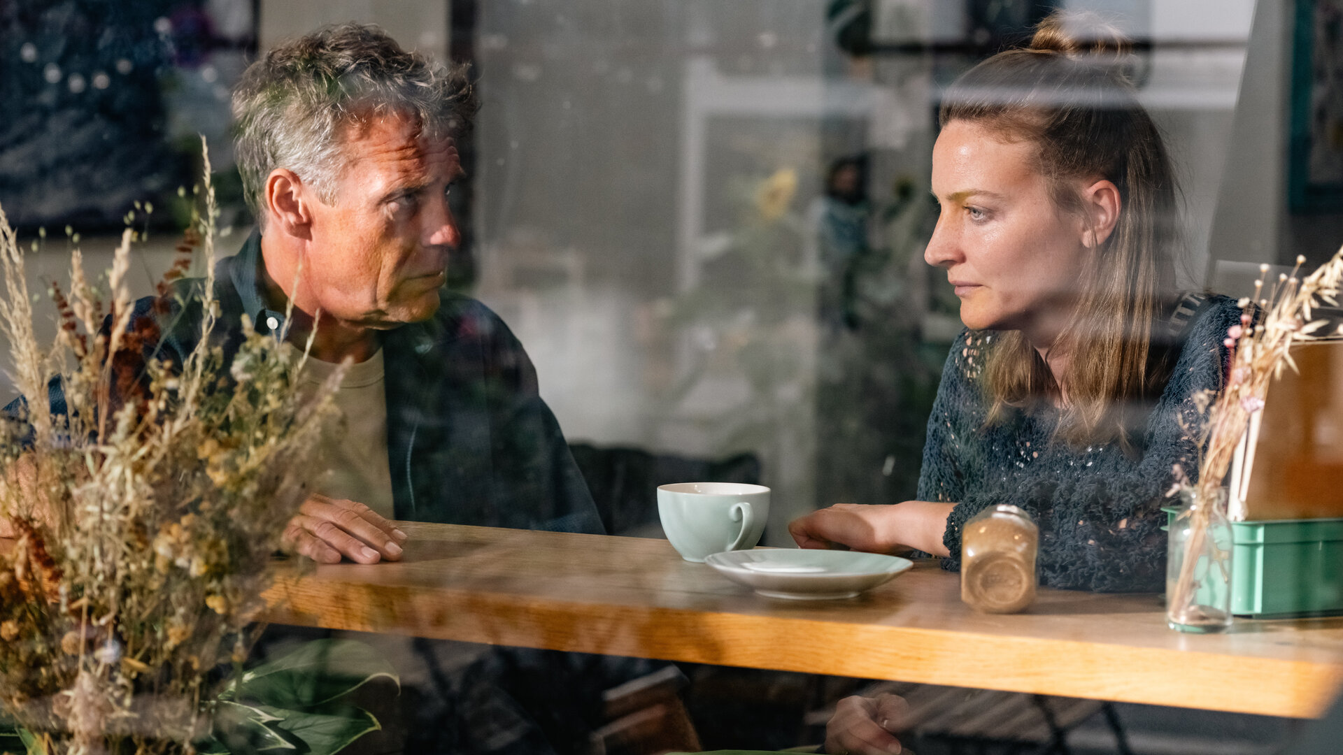 "SOKO Köln - Ungebunden": Susann Mayrath (Ines Marie Westernströer) sitzt neben Matti Wagner (Pierre Besson) hinter einer Fensterscheibe in einem Café an einem hohen Tisch. Vor ihr auf dem Tisch steht ein leerer Teller und eine Tasse. Sie blickt traurig nach vorne. Matti sieht sie ernst an. Rechts im Bildrand steht hinter der Scheibe ein Strauß Trockenblumen.