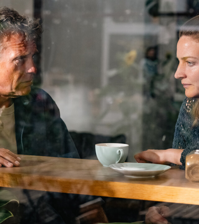 "SOKO Köln - Ungebunden": Susann Mayrath (Ines Marie Westernströer) sitzt neben Matti Wagner (Pierre Besson) hinter einer Fensterscheibe in einem Café an einem hohen Tisch. Vor ihr auf dem Tisch steht ein leerer Teller und eine Tasse. Sie blickt traurig nach vorne. Matti sieht sie ernst an. Rechts im Bildrand steht hinter der Scheibe ein Strauß Trockenblumen.