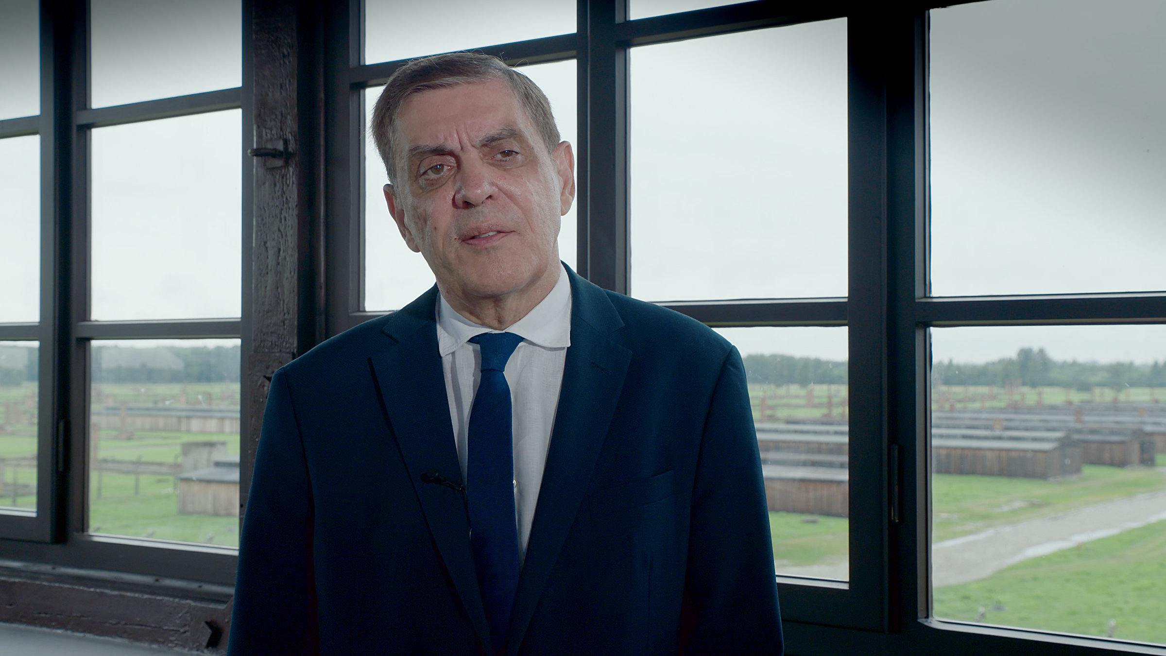 „Unrecht und Widerstand - Romani Rose und die Bürgerrechtsbewegung“:  Ein älterer bartloser Mann mit graumelierten kurzen Haaren, der ein dunkelblaues Sakko und einen dunkelblauen Schlips und ein weißes Hemd trägt – Romani Rose -, steht vor einem Fenster im ersten oder zweiten Stock eines Gebäudes und spricht. Hinter ihm hat man die Aussicht über die Überreste des ehemaligen Lagers Auschwitz-Birkenau.