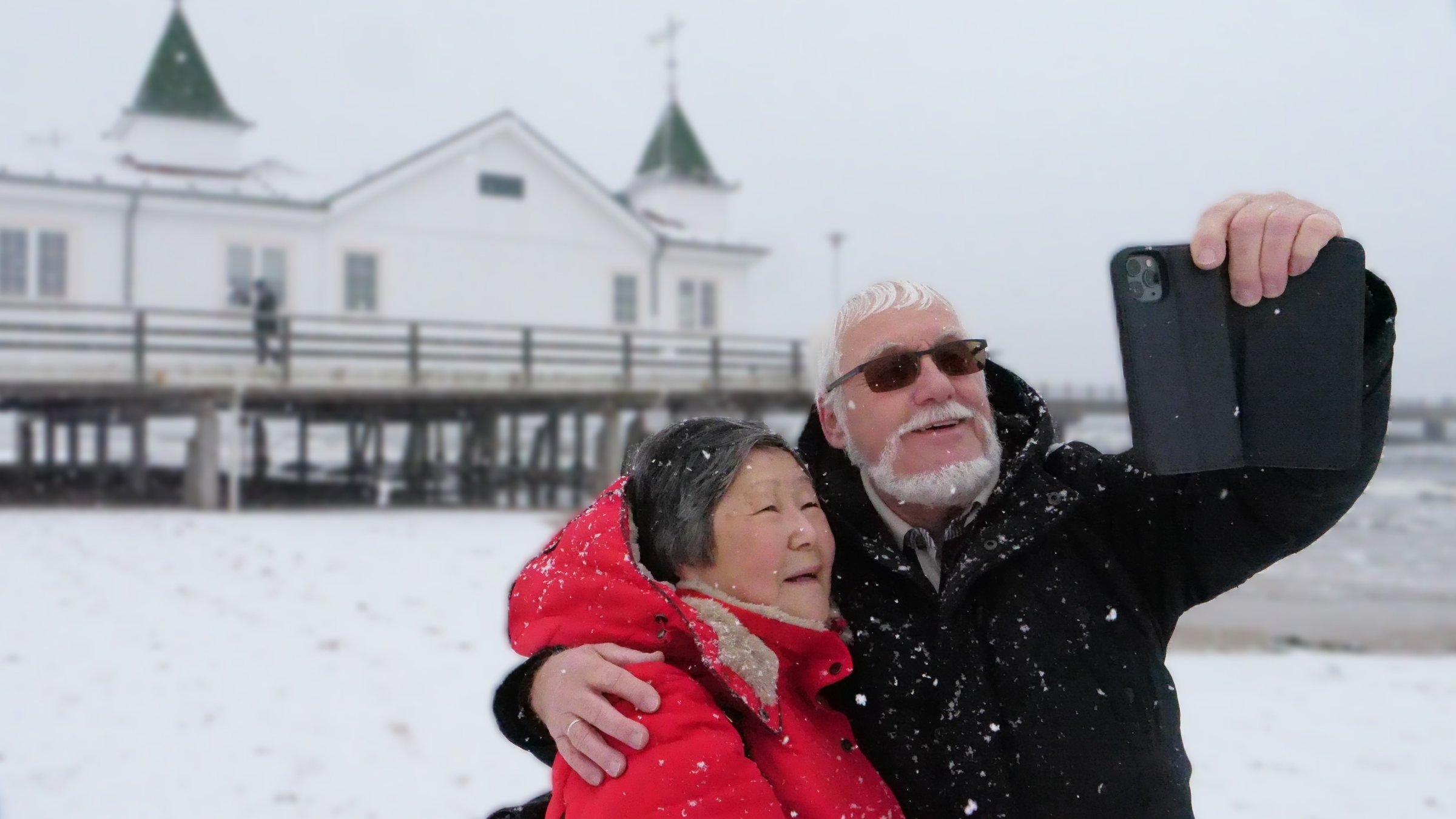 "ZDF.reportage: Usedom im Winter - Auszeit auf der Insel": Ein Paar, ein weißhariger Mann mit Bart und eine Frau mit roter Jacke, stehen vor der Ahlbecker Seebrücke im Schneetreiben und machen mit dem Smartphone ein Foto von sich selbst.