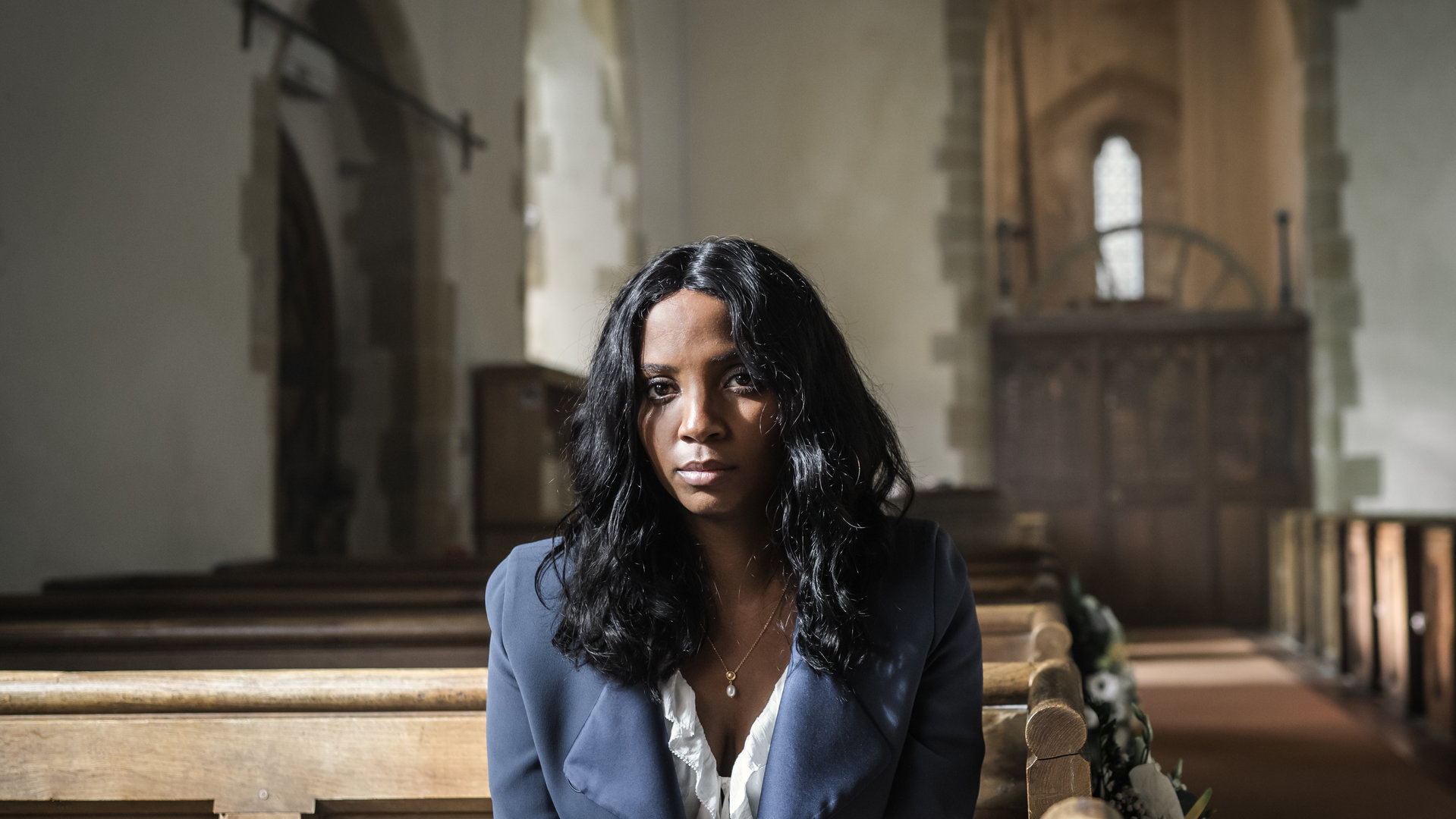 "Grace - Stirb ewig": Ashley Haynes (Alisha Bailey) sitzt in einer leeren Kirche auf einer Bank und schaut mit leerem Blick nach vorne. Sie trägt ein Brautkleid und darüber eine blaue Jacke.