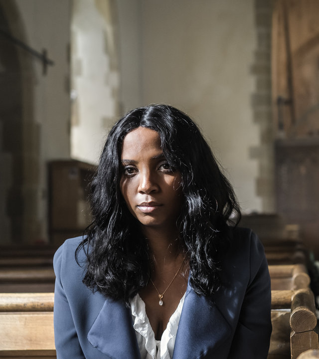 "Grace - Stirb ewig": Ashley Haynes (Alisha Bailey) sitzt in einer leeren Kirche auf einer Bank und schaut mit leerem Blick nach vorne. Sie trägt ein Brautkleid und darüber eine blaue Jacke.