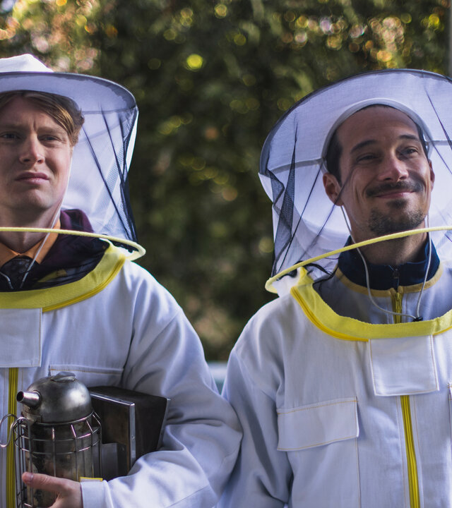 "SPYON - Der erste Außeneinsatz": Asel Asanow (Lukas Zumbrock) und Andreas Rödig (Klaus Steinbacher) stehen im Freien tragen Imkeranzüge. Asel Asanow hält einen Bienen-Smoker in der Hand.