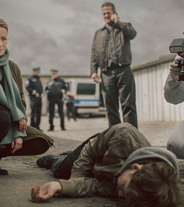 "Theresa Wolff - Nebel": Auf einem Garagenhof liegt Finn Behrend (Philip Günsch) tot, seitlich auf dem Boden. Dr. Theresa Wolff (Nina Gummich) kniet daneben während eine Mitarbeiterin der Spurensicherung (Komparsin) die Leiche fotografisch dokumentiert. Im Hintergrund telefoniert Bruno Lewandowski (Aurel Manthei) mit seinem Handy.