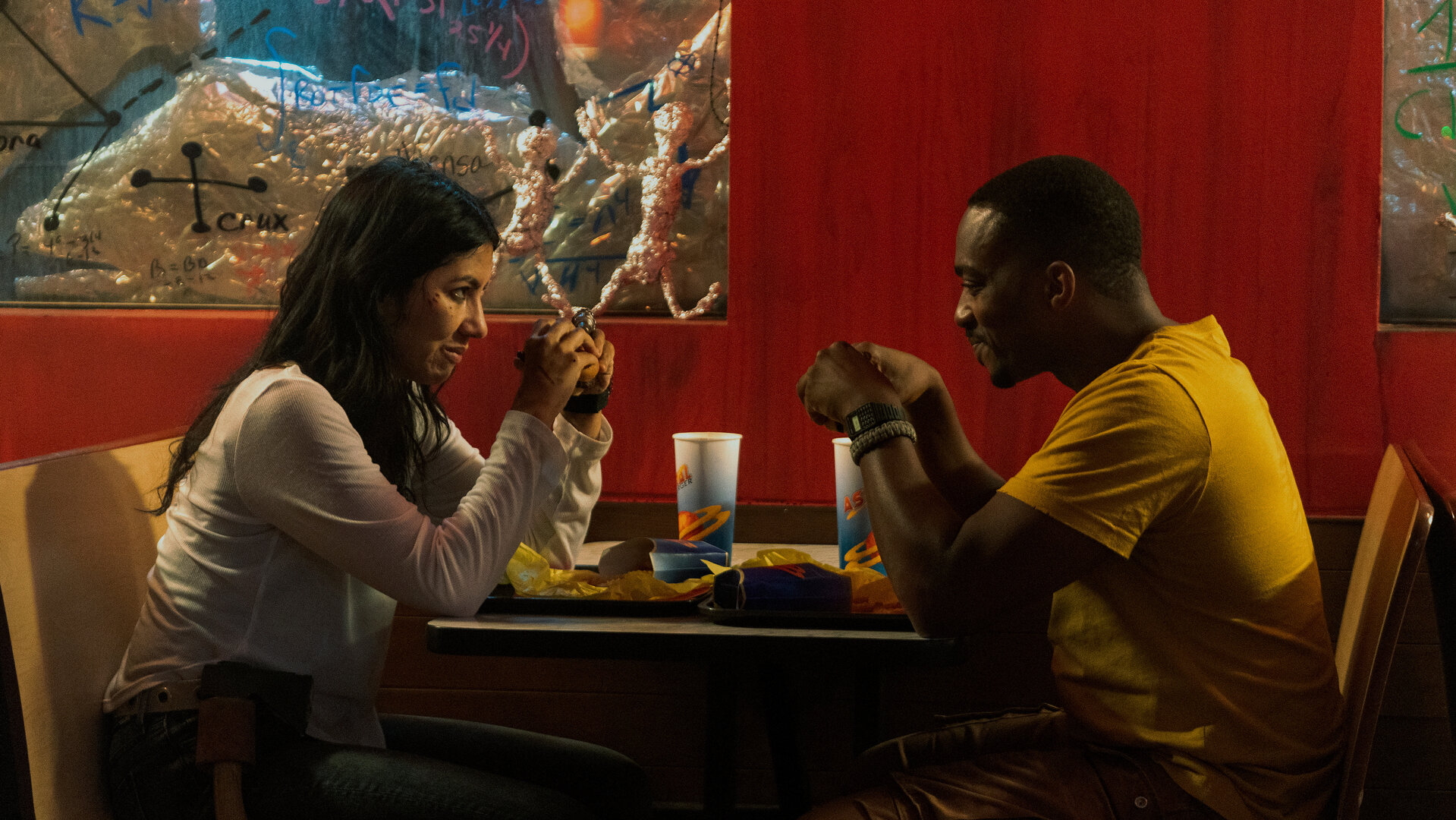 "Twisted Metal - DRVTHRU": John (Anthony Mackie) und Quiet (Stephanie Beatriz) sitzen sich an einem Tisch in einem Diner gegenüber und blicken sich lächelnd an. Vor ihnen stehen Tabletts mit Essen und Getränkebechern, im Hintergrund stehen Figuren aus Alufolie und auf den Fensterscheiben sind handgeschriebene Formeln.