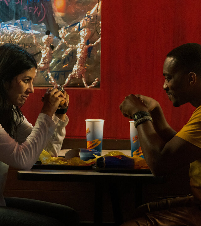 "Twisted Metal - DRVTHRU": John (Anthony Mackie) und Quiet (Stephanie Beatriz) sitzen sich an einem Tisch in einem Diner gegenüber und blicken sich lächelnd an. Vor ihnen stehen Tabletts mit Essen und Getränkebechern, im Hintergrund stehen Figuren aus Alufolie und auf den Fensterscheiben sind handgeschriebene Formeln.