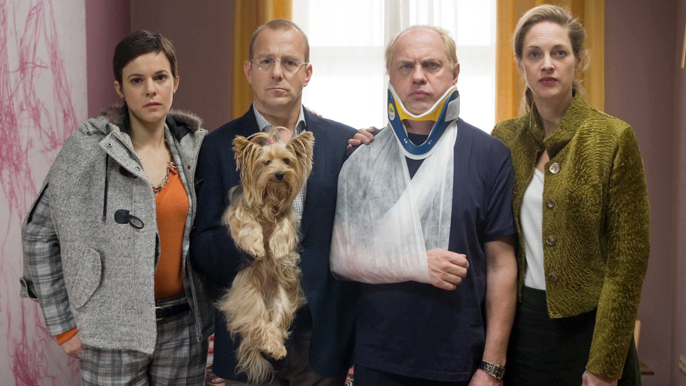 "Ein vorbildliches Ehepaar": Vicky Berger (Annika Kuhl), Roman Ellermann (Heino Ferch) mit einem Yorkshire Terrier auf dem Arm, Thomas Berger (Uwe Ochsenknecht) mit Halskrause und Arm in einer Schlinge und Isabelle Ellermann (Sophie von Kessel) stehen nebeneinander in einem Wartezimmer.