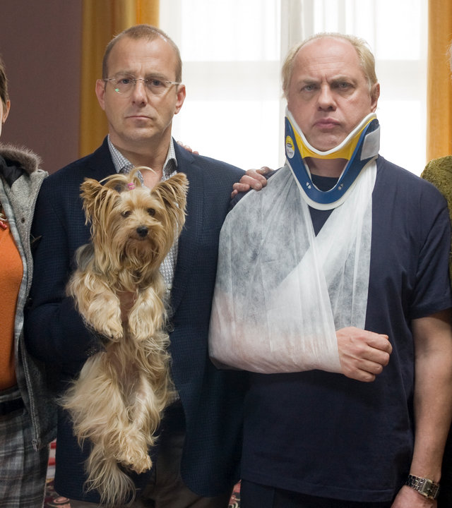 "Ein vorbildliches Ehepaar": Vicky Berger (Annika Kuhl), Roman Ellermann (Heino Ferch) mit einem Yorkshire Terrier auf dem Arm, Thomas Berger (Uwe Ochsenknecht) mit Halskrause und Arm in einer Schlinge und Isabelle Ellermann (Sophie von Kessel) stehen nebeneinander in einem Wartezimmer.