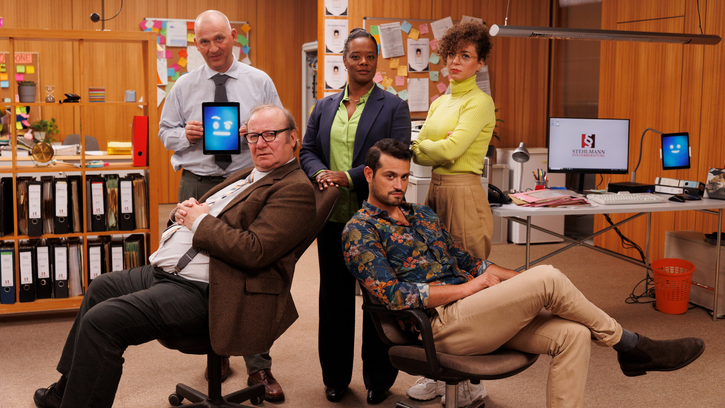 "Browser Ballett - KI-Kollege": In einem Büro mit holzvertäfelten Wänden präsentiert sich ein Teil des Teams von "Stehlmann Steuerberatung". Auf zwei Schreibtischstühlen sitzen vorne Volker (Rainer Reiners) und Ben (Nicolas F. Türksever), hinter ihnen stehen ihre Kolleg:innen Arne (Steffen Roll) und Pia (Christina Schlag), sowie ihre Chefin Eva (Denise M'Baye). Arne hält ein Tablet in der Hand, das einen skeptisch schauenden Smiley zeigt. Es ist der KI-Kollege.