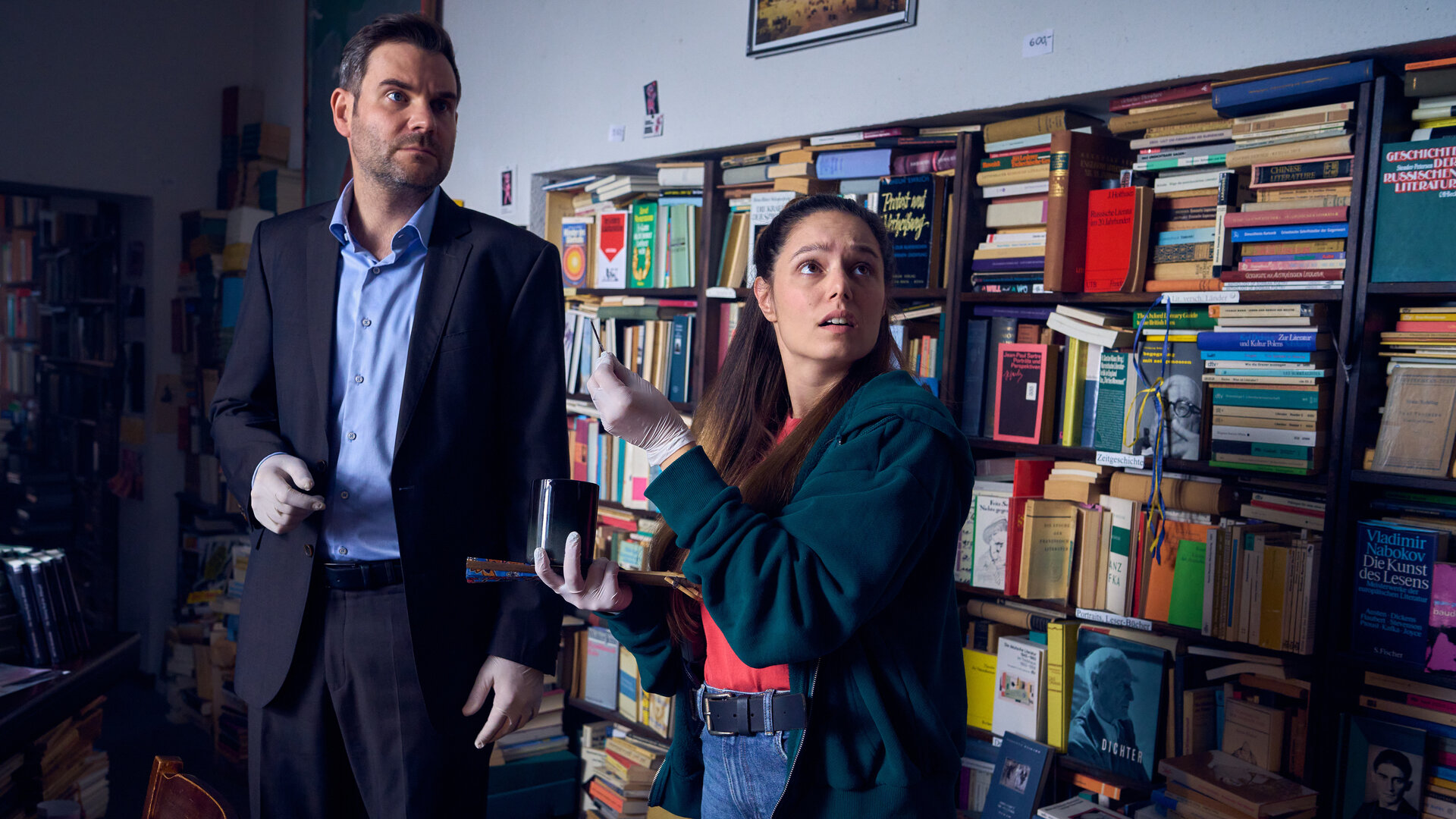 "SOKO Köln - Der verfluchte Jonas": Jonas Fischer (Lukas Piloty) und Amira Mahdi (Aylin Ravanyar) stehen nebeneinander im Antiquariat vor einem großen Bücherregal und sehen ernst in die gleiche Richtung. Amira hat den Mund erstaunt geöffnet. Sie hält ein dunkles Gefäß und ein schwarzes Stäbchen in ihren behandschuhten Händen. Jonas trägt ebenfalls weiße Gummihandschuhe.