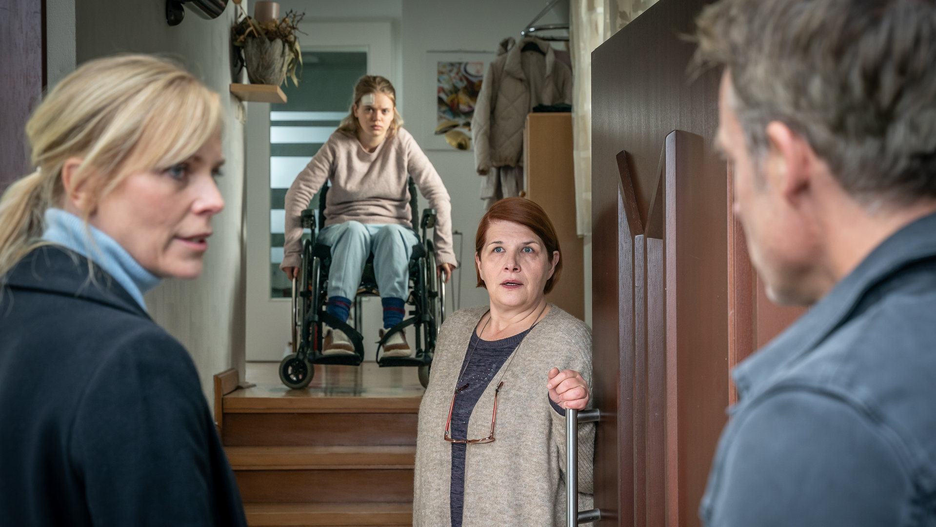 "SOKO Leipzig - Die heilige Agnes": Ina Zimmermann (Melanie Marschke) und Jan Maybach (Marco Girnth) stehen vor der Haustür von Dorit Engelmann (Susanne Böwe), die gerade die Tür öffnet. Im Hintergrund sitzt Greta Engelmann (Maria Matschke Engel) auf einem Treppenabsatz im Rollstuhl. Sie hat ein Pflaster auf der Stirn. Ina sieht Jan fragend an.