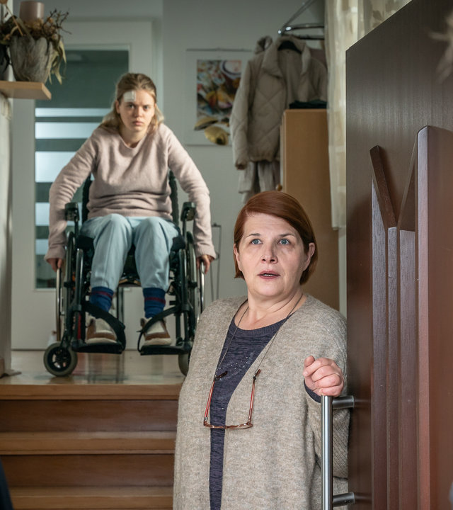 "SOKO Leipzig - Die heilige Agnes": Ina Zimmermann (Melanie Marschke) und Jan Maybach (Marco Girnth) stehen vor der Haustür von Dorit Engelmann (Susanne Böwe), die gerade die Tür öffnet. Im Hintergrund sitzt Greta Engelmann (Maria Matschke Engel) auf einem Treppenabsatz im Rollstuhl. Sie hat ein Pflaster auf der Stirn. Ina sieht Jan fragend an.
