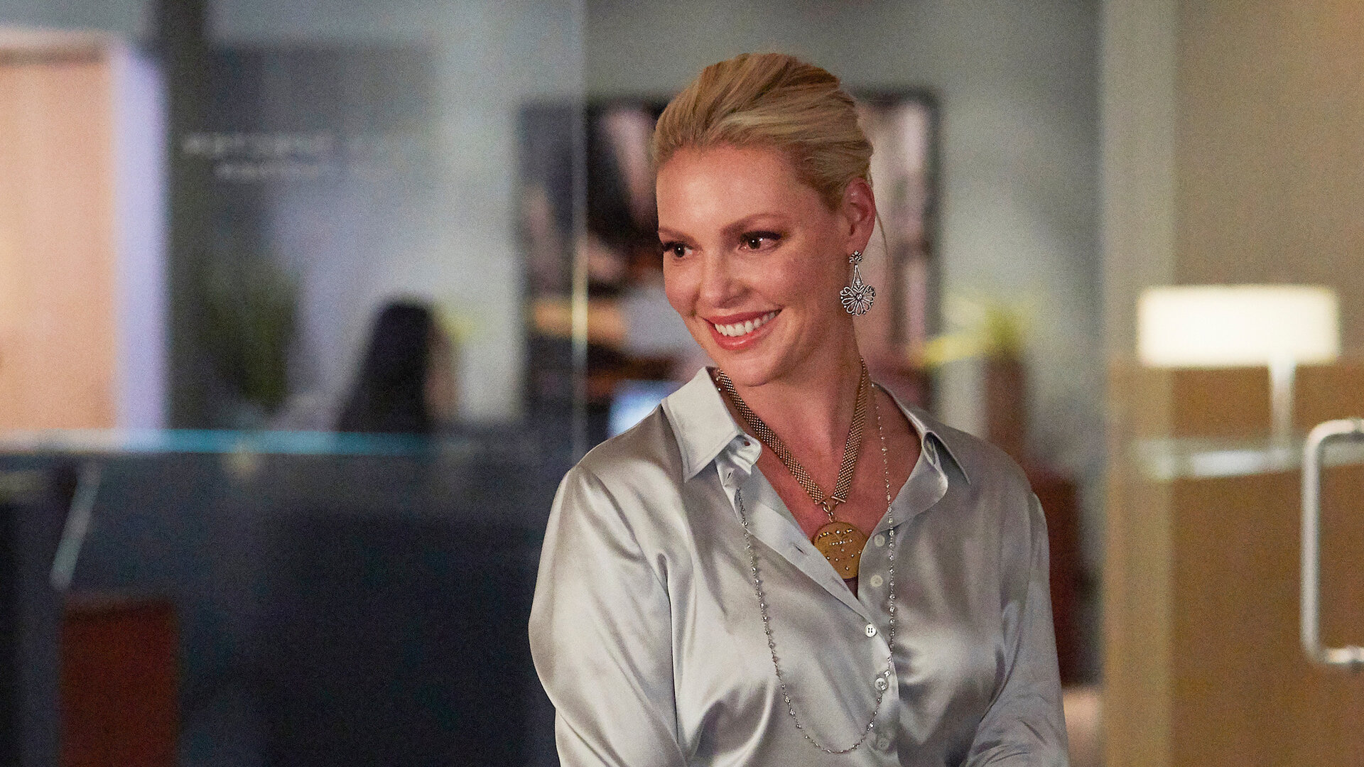 "Suits - Rangordnung": Im monochromen, silberglänzenden Look sitzt die lächelnde Samantha (Katherina Heigl) auf der Lehne eines Sessels. Im Hintergrund ist durch die Glasfront des Büros der Flurbereich der Kanzlei zu erkennen, wo sich drei weitere Mitarbeiter aufhalten.