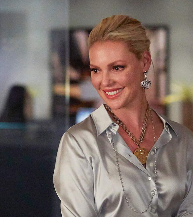 "Suits - Rangordnung": Im monochromen, silberglänzenden Look sitzt die lächelnde Samantha (Katherina Heigl) auf der Lehne eines Sessels. Im Hintergrund ist durch die Glasfront des Büros der Flurbereich der Kanzlei zu erkennen, wo sich drei weitere Mitarbeiter aufhalten.