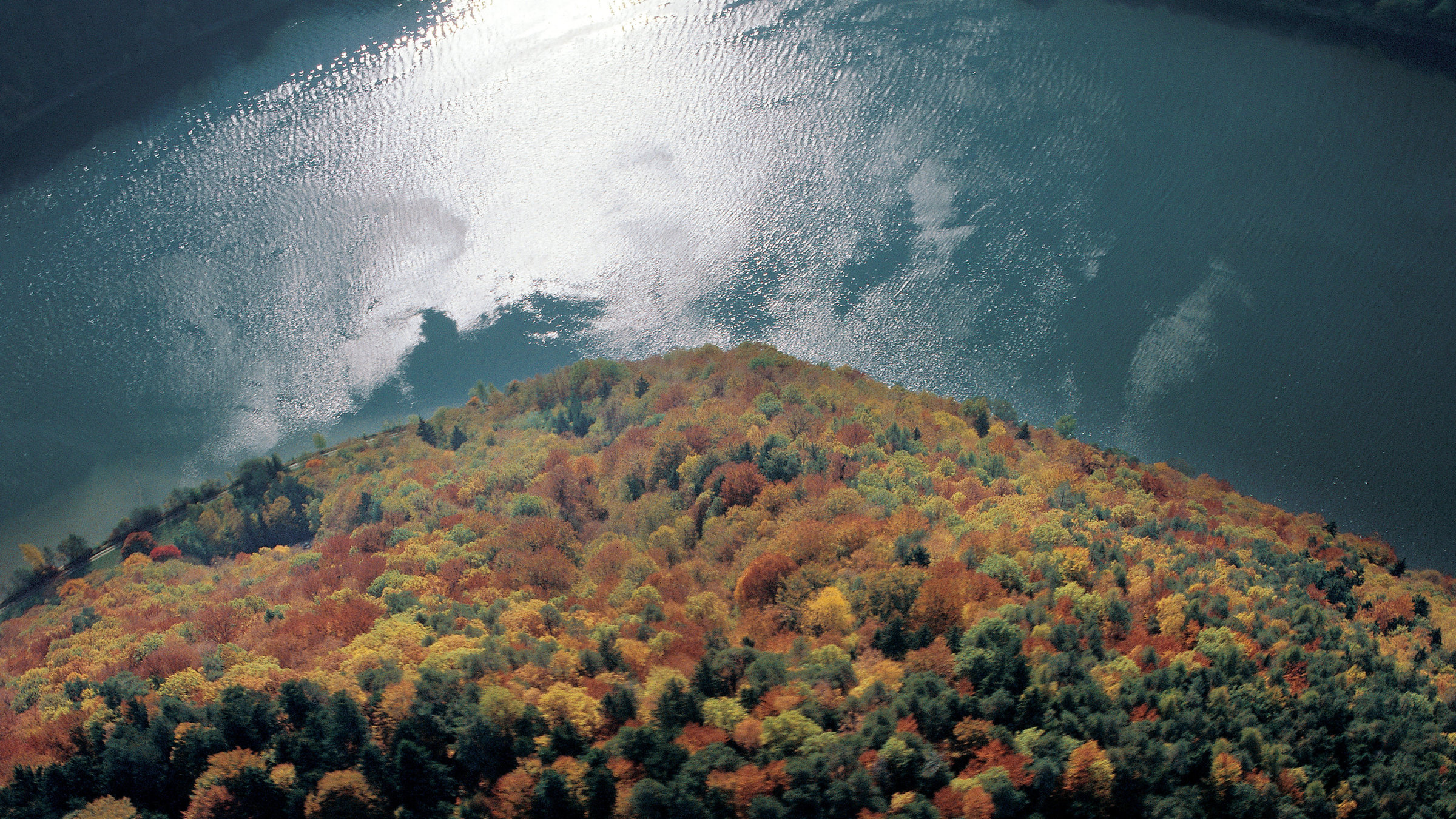 "Über Österreich - Die dritte Erkundung - Der Norden": Blick von oben auf einen bewaldeten Berg, dahinter ein See