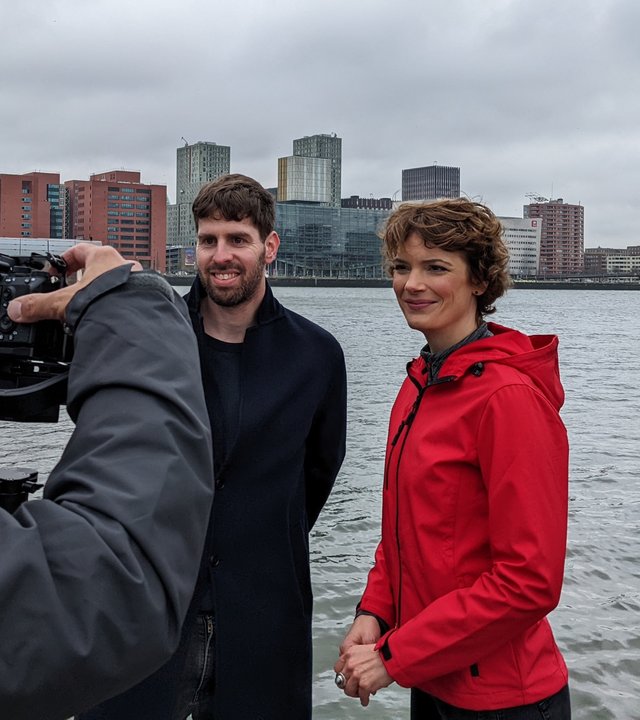 Lior Steinberg und Lena Ganschow stehen am Flussufer und schauen lächelnd in eine Kamera. Der Kameramann ist im Vordergrund links im Anschnitt zu sehen. Im Hintergrund sieht man Häuser in Rotterdam.