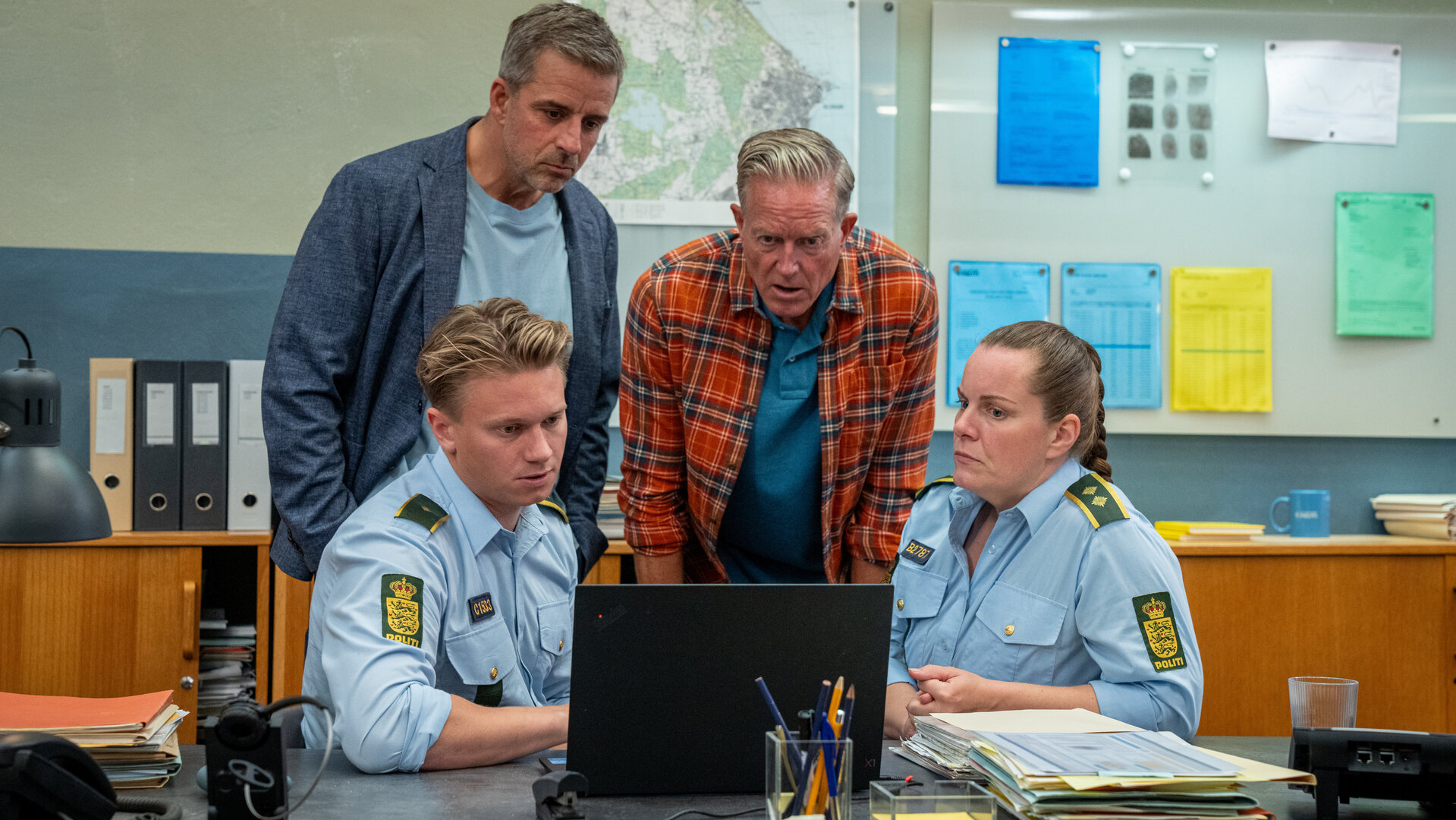 "Dan Sommerdahl - Tödliche Idylle: Die Enttäuschung": Benjamin (Mathias Käki Jørgensen, l.) sitzt an seinem Schreibtisch vor einem Laptop, auf dem er Flemming (André Babikian, 2. v. l.) und Dan (Peter Mygind), die hinter ihm stehen, sowie Nadia (Lise Baastrup), die neben ihm sitzt, etwas zeigt.