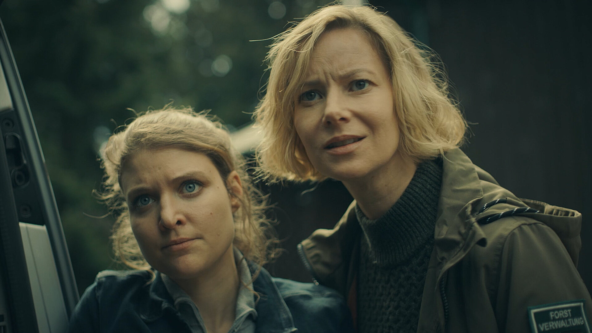 "Erzgebirgskrimi – Über die Grenze": Karina Szabo (Lara Mandoki) und Saskia Bergelt (Teresa Weißbach) stehen nah beieinander an einer Autotür. Sie schauen mit angespanntem Gesichtsausdruck seitlich an der Kamera vorbei.