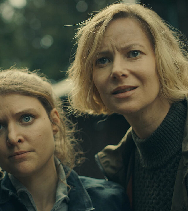 "Erzgebirgskrimi – Über die Grenze": Karina Szabo (Lara Mandoki) und Saskia Bergelt (Teresa Weißbach) stehen nah beieinander an einer Autotür. Sie schauen mit angespanntem Gesichtsausdruck seitlich an der Kamera vorbei.