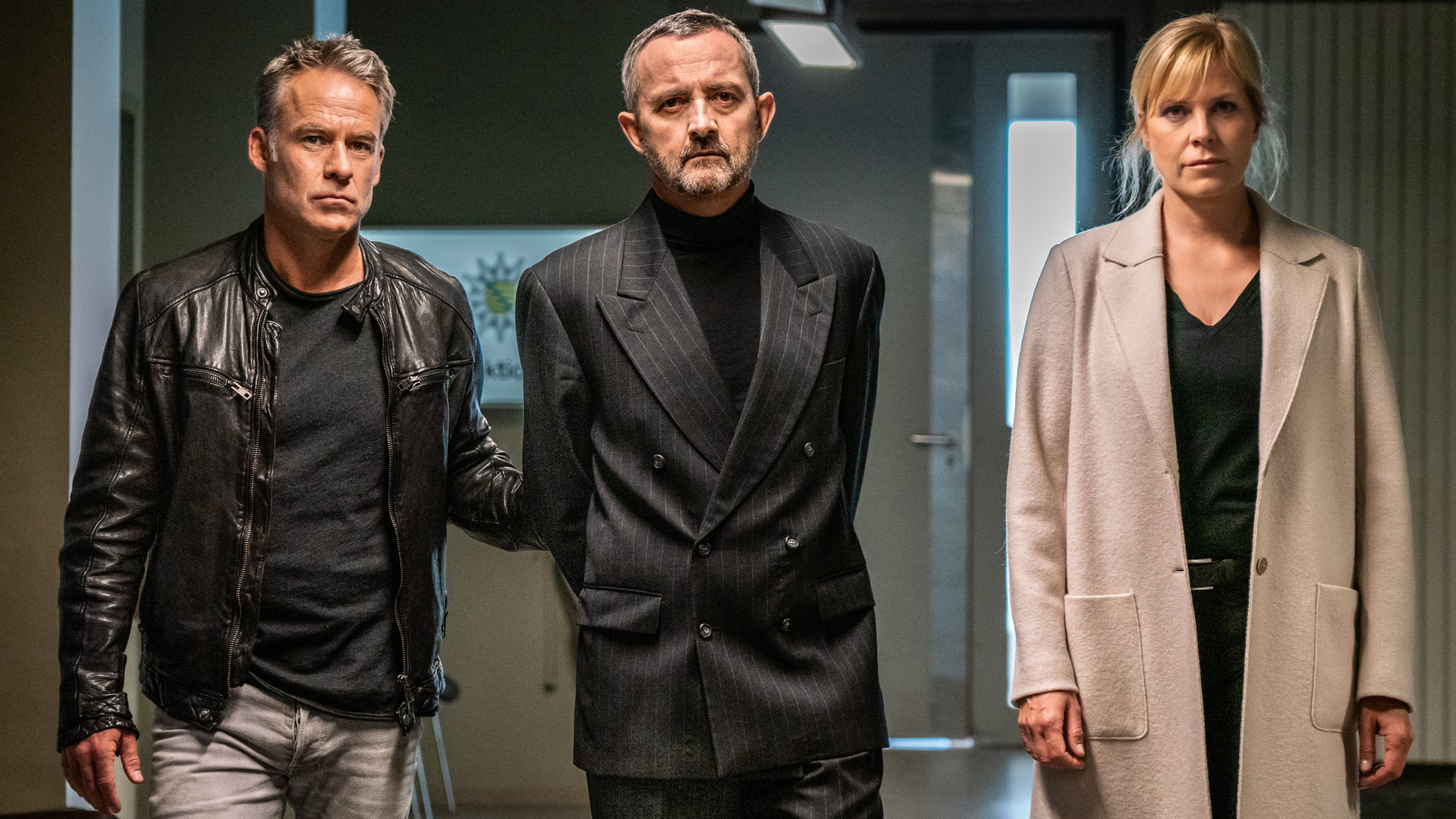 "SOKO Leipzig - Family Business (2)": Jan Maybach (Marco Girnth), Davide Di Maria (Harald Schrott) und Ina Zimmermann (Melanie Marschke) auf dem Gang der Polizeidirektion. Davide hat die Hände auf dem Rücken fixiert und wird von Jan und Ina abgeführt.