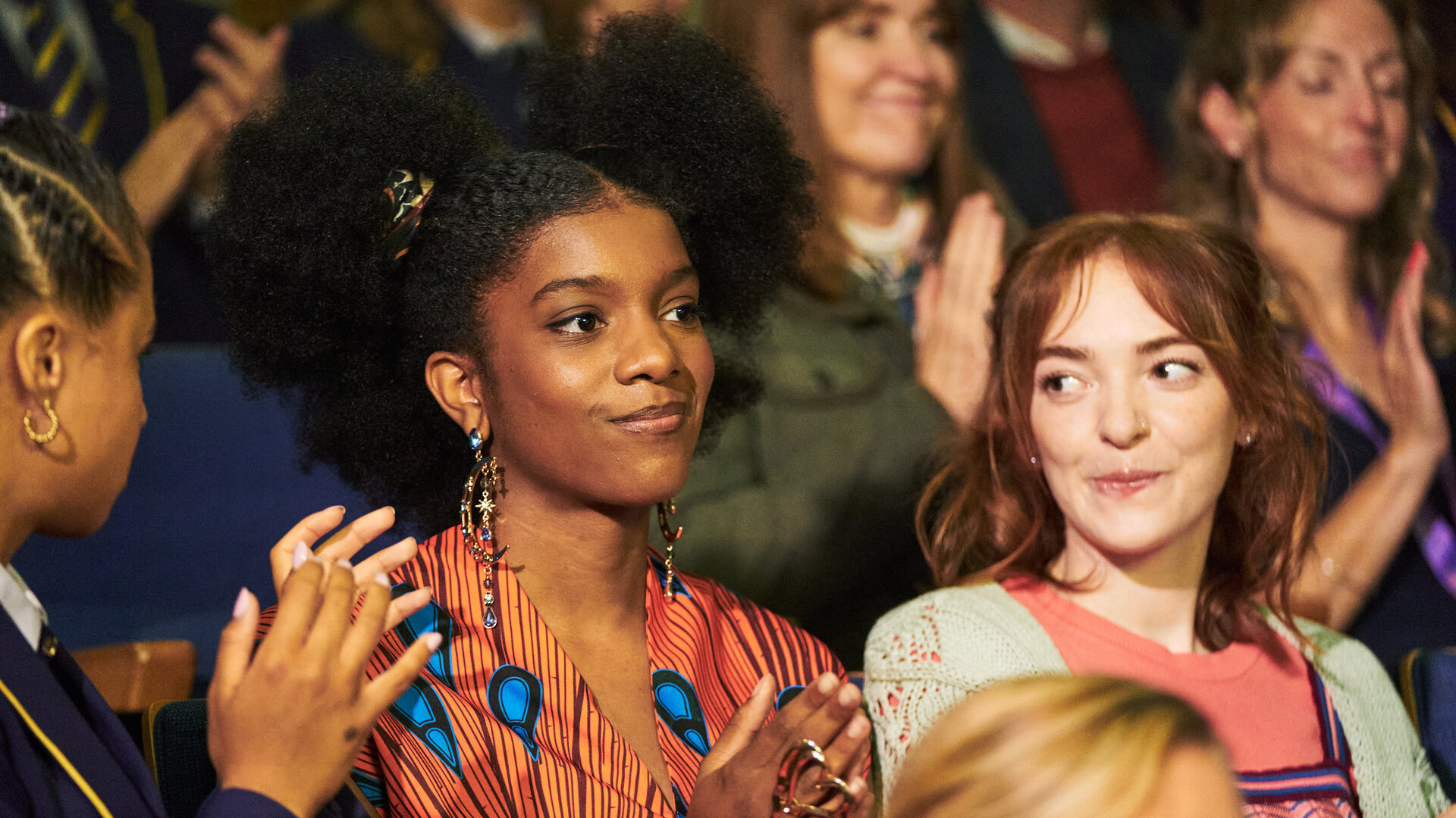 "Boarders - Welcome to St. Gilbert's - Money & Monologues": Abby (Assa Kanouté), Devonyé (Kendra Brown) und Mabel (Georgina Sadler) sitzen in einer Gruppe anderer Schülerinnen (Komparsinnen) und applaudieren. Abby und Mabel schauen Devonyé an, die selbstbewusst nach vorne blickt.