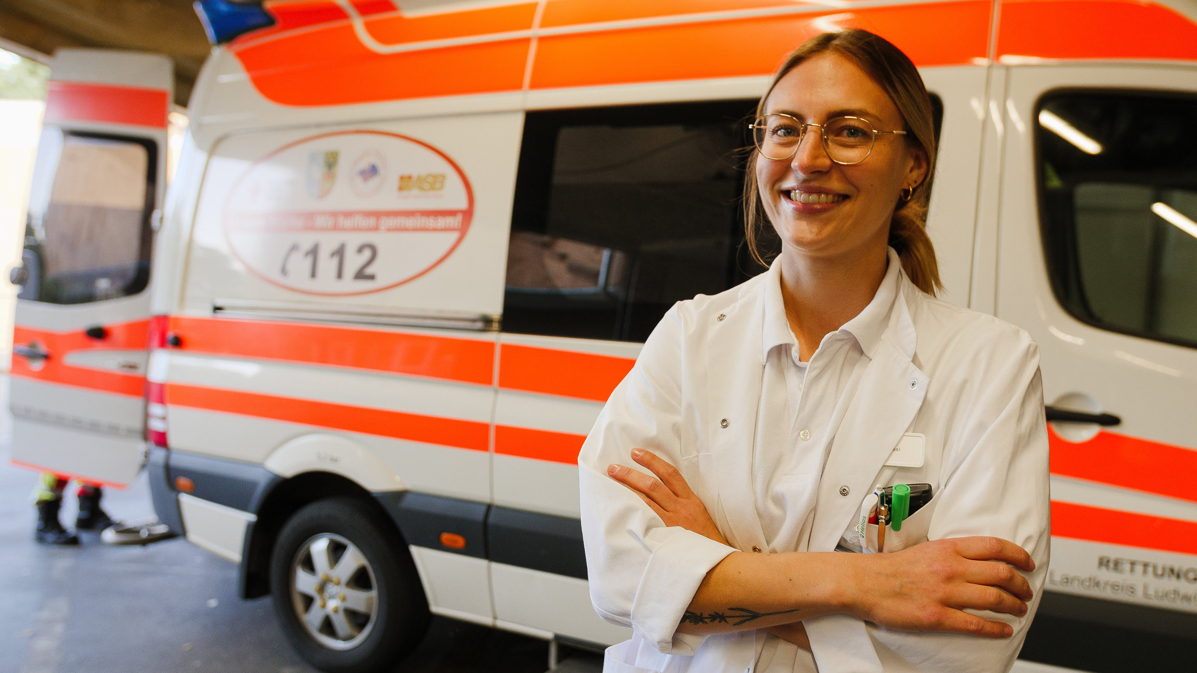 "37 Grad: Dienste, Druck und Dauerstress - Junge Docs in der Klinik": Assistenzärztin Jule (26) steht vor einem Rettungswagen und lächelt in die Kamera. Sie trägt einen weißen Arztkittel, eine Brille und ihre blonden langen Haare zu einem Zopf.