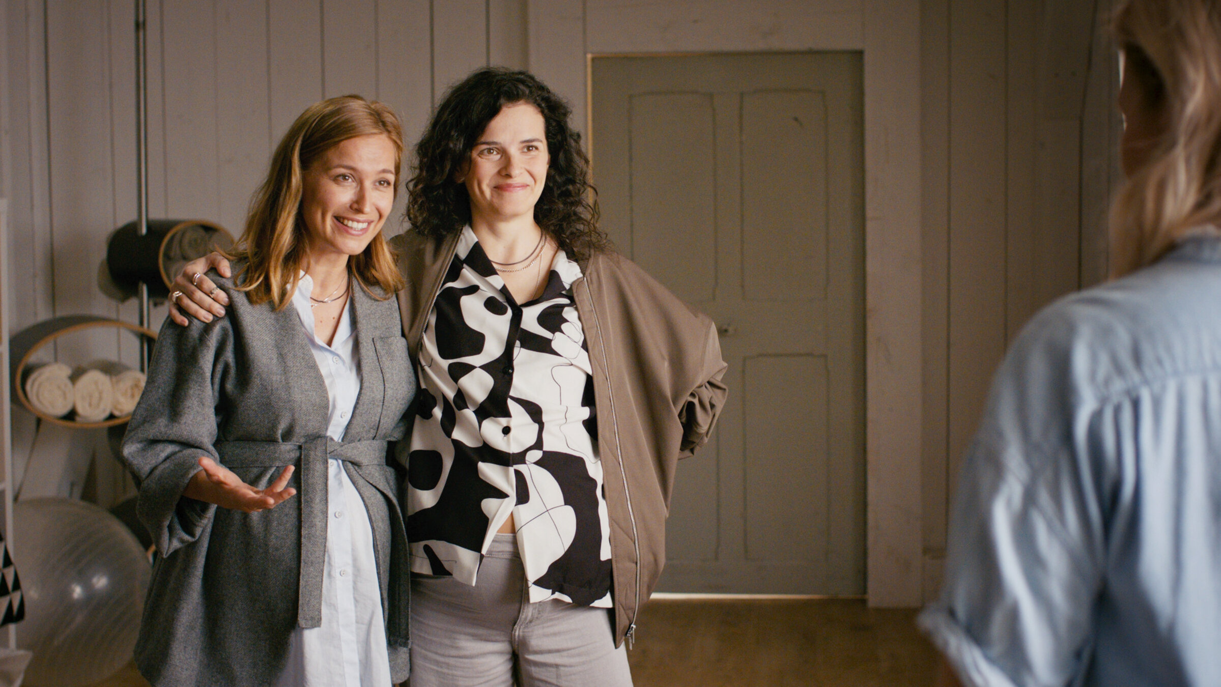 "Lena Lorenz - Kreuz und queer": In der Hebammenpraxis stehen Alex (Pina Kühr) und Franzi Winkler-Ertl (Maxine Kazis) gegenüber von Lena Lorenz (Judith Hoersch), die mit dem Rücken zur Kamera steht. Die beiden Frauen haben die Arme umeinander gelegt und schauen Lena lachend an.