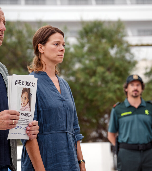 "Lillys Verschwinden (1)": Robert Bischoff (Heino Ferch) und Anna Bischoff (Jessica Schwarz) stehen mit einem Plakat ihrer vermissten Tochter auf der Straße nebeneinander. Zwei Polizisten (Komparsen) stehen im Hintergrund.