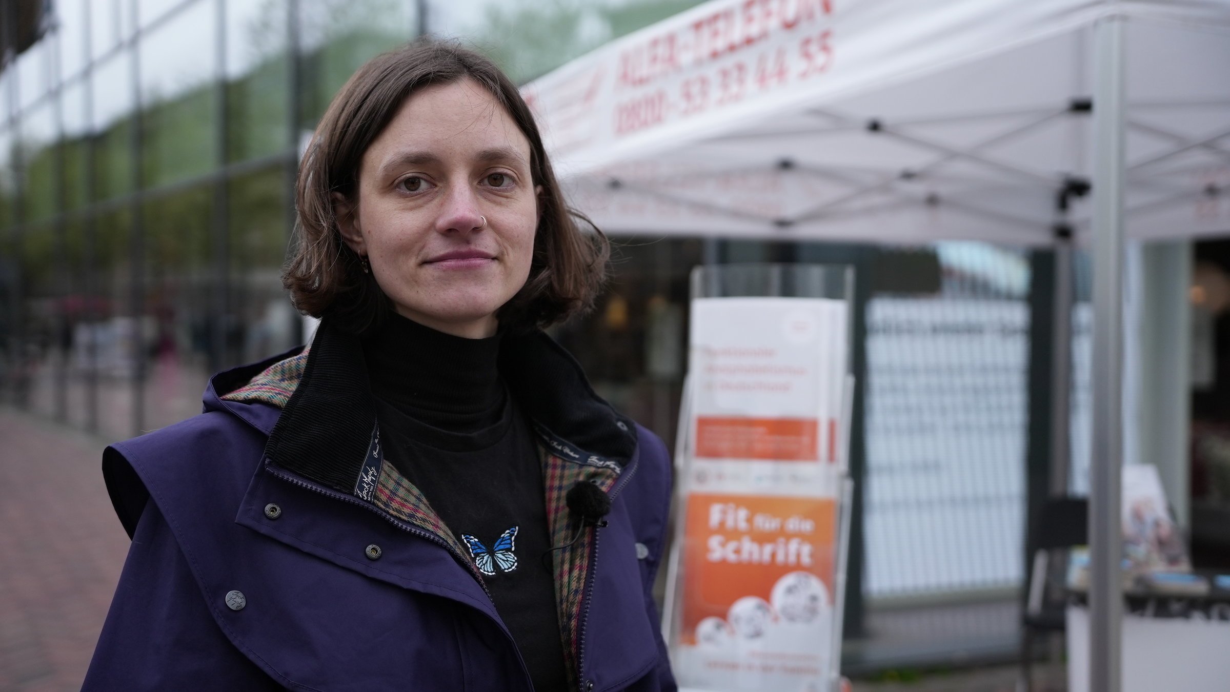 "37°: Immer Ärger mit den Buchstaben - Wenn das Alphabet zur Qual wird": Kunststudentin Lea steht vor einem Infostand, auf dem Hilfsangebote für Menschen, die nicht richtig lesen und schreiben können, beworben werden. Die Frau schaut selbstbewusst in die Kamera.