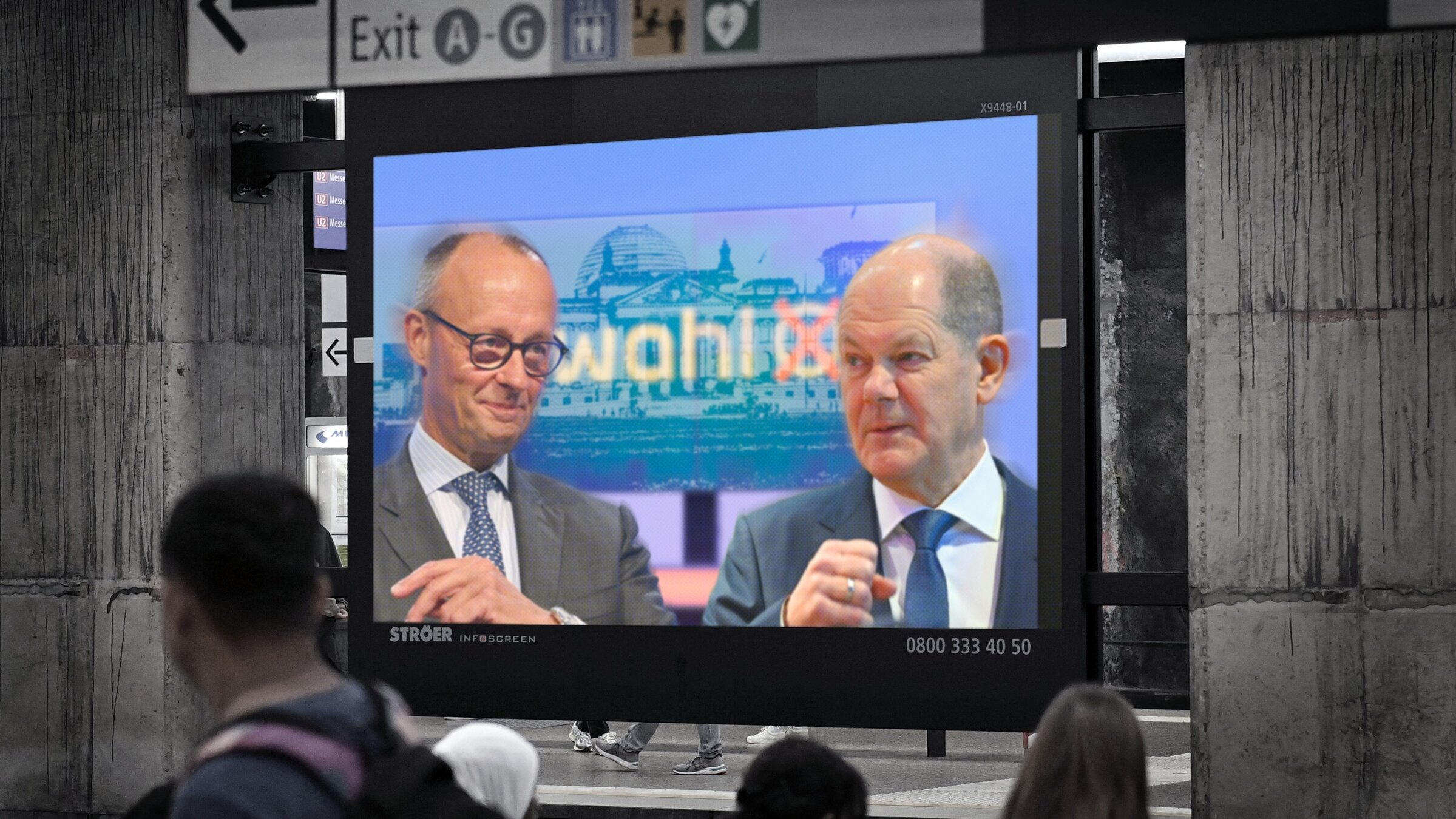 "Kanzler und Herausforderer - Scholz und Merz im Wahlkampf": Fotomontage: Auf einem Bildschirm in einer U-Bahnstation sind Friedrich Merz und Olaf Scholz zu sehen. Im Vordergrund sind Hinterköpfe von Menschen, im Hintergrund über dem Bildschirm befinden sich Wegweiser.