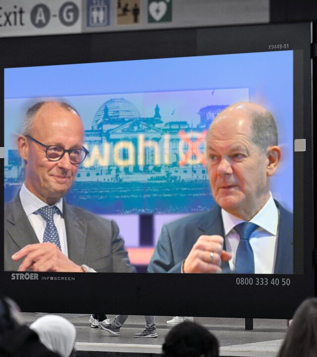 "Kanzler und Herausforderer - Scholz und Merz im Wahlkampf": Fotomontage: Auf einem Bildschirm in einer U-Bahnstation sind Friedrich Merz und Olaf Scholz zu sehen. Im Vordergrund sind Hinterköpfe von Menschen, im Hintergrund über dem Bildschirm befinden sich Wegweiser.