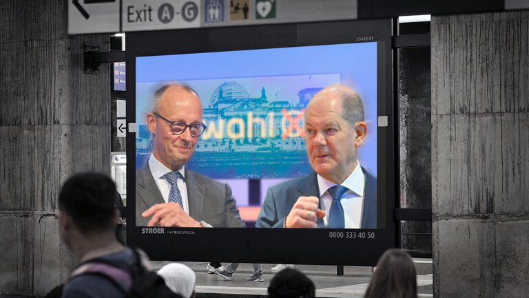 "Kanzler und Herausforderer - Scholz und Merz im Wahlkampf": Fotomontage: Auf einem Bildschirm in einer U-Bahnstation sind Friedrich Merz und Olaf Scholz zu sehen. Im Vordergrund sind Hinterköpfe von Menschen, im Hintergrund über dem Bildschirm befinden sich Wegweiser.