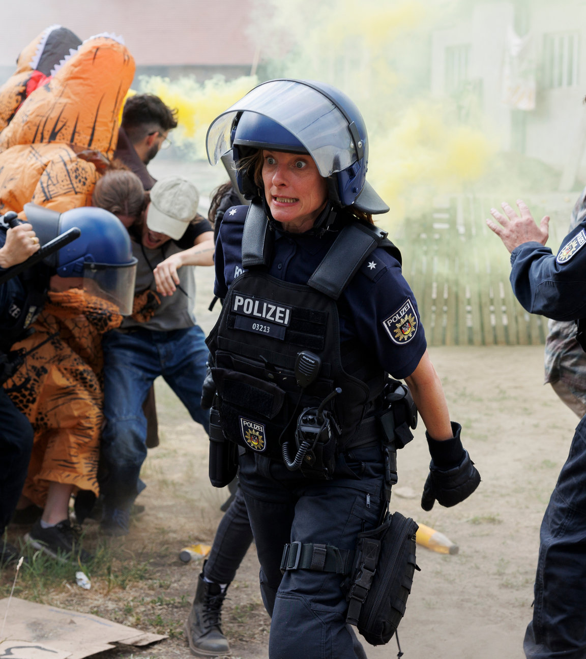 "Browser Ballett: Fire for Future": Während schwer ausgerüstete Polizisten mit – teils verkleideten – Demonstrant:innen aufeinander und zusammenstoßen, ist Einsatzleiterin Anja Klopp (Anne Ratte-Polle) anzusehen, dass ihr die Situation entgleitet. Sie schaut panisch. Im Hintergrund sind bunte Rauchschwaden zu sehen.