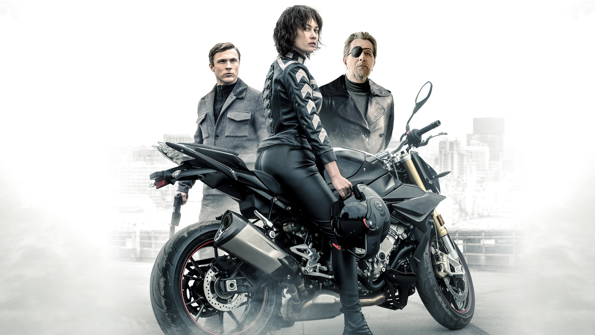 "The Courier - Tödlicher Auftrag": Der Kurier (Olga Kurylenko) sitzt auf einem Motorrad und sieht zur Kamera. Hinter dem Kurier sind Ezekiel Mannings (Gary Oldman) und Agent Bryant (William Moseley) zu sehen.