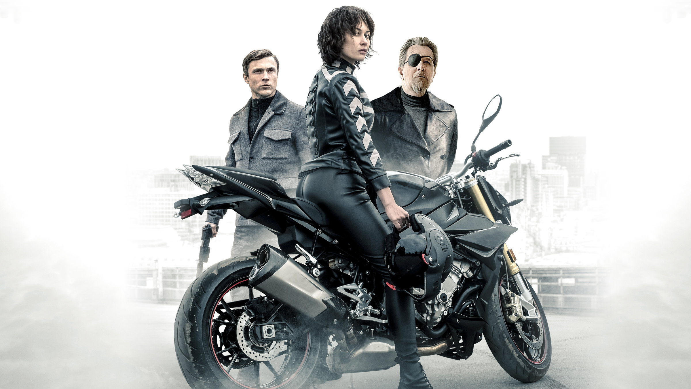 "The Courier - Tödlicher Auftrag": Der Kurier (Olga Kurylenko) sitzt auf einem Motorrad und sieht zur Kamera. Hinter dem Kurier sind Ezekiel Mannings (Gary Oldman) und Agent Bryant (William Moseley) zu sehen.