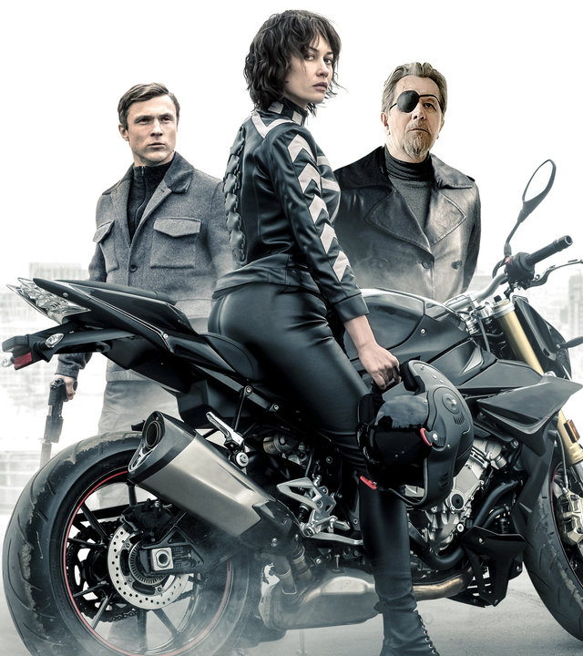 "The Courier - Tödlicher Auftrag": Der Kurier (Olga Kurylenko) sitzt auf einem Motorrad und sieht zur Kamera. Hinter dem Kurier sind Ezekiel Mannings (Gary Oldman) und Agent Bryant (William Moseley) zu sehen.