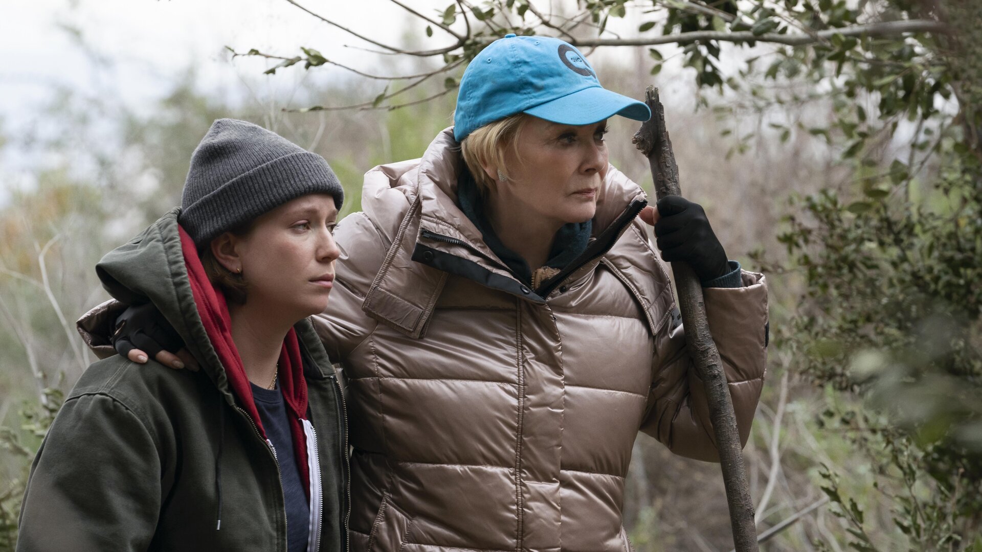 "Hacks - Eines Tages ist jetzt": Deborah und Ava stehen in Outdoorkleidung nebeneinander. Deborah hält in ihrer Hand einen großen Stock, auf den sie sich zu stützt. Um sie herum stehen Bäume und Sträucher.