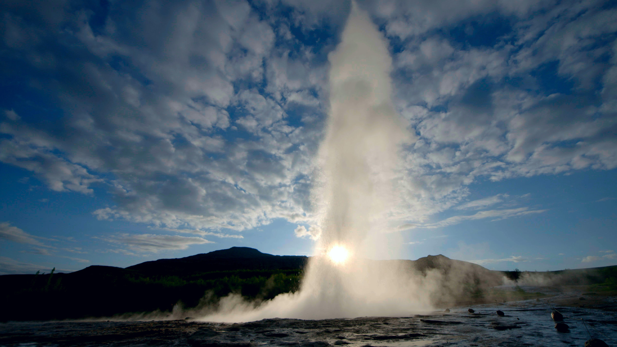 "Island - Weltspitze": Drei Viertel des Bildes nimmt ein schwach bewölkter Himmel ein, wobei die Sonne mittig knapp oberhalb des Horizonts liegt. Dieser wird von einem baumlosen flach ansteigenden grünen Hügel gebildet. Zentraler Mittelpunkt des Bildes ist eine hohe Wasserfontäne, entstanden durch die Eruption eines Geysirs.
