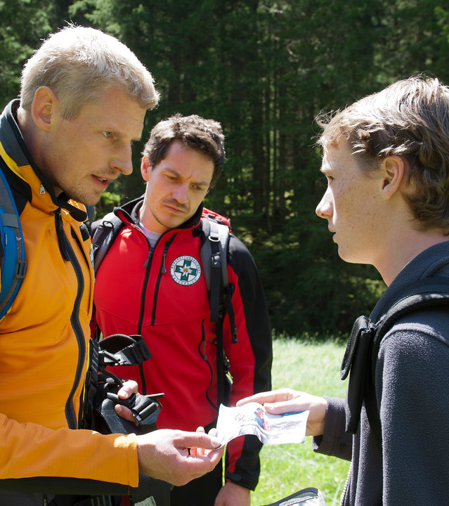 "Die Bergretter: Wilde Wasser - Teil 2": Andreas (Martin Gruber) steht Timo (Simon Jung) auf einer Waldlichtung gegenüber. Dieser hält eine Geldbörse in der Hand und zeigt Andreas ein Foto. Tobias (Markus Brandl) steht neben ihnen und blickt ebenfalls auf das Foto.