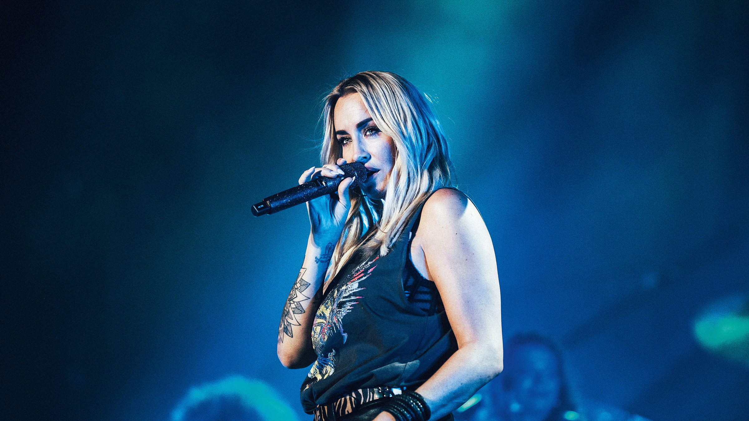"Sarah Connor: Herz Kraft Werke - Live": Eine Frau mit langen blonden Haaren steht vor einem blau beleuchteten Hintergrund auf der Bühne. Sie hält das Mikrofon dicht vor ihrem Mund und schaut lasziv in die Kamera.